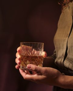 Close-up of a hand holding a cut-glass tumbler filled with amber whiskey against a dark background.